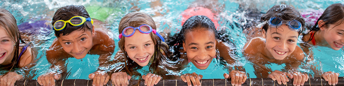 Children in the water lined up on the edge of the pool smiling up at the camera